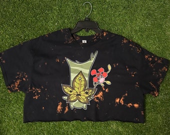 May include: Black crop top with a cartoon graphic of a plant with a red flower. The shirt has a bleached design and is hanging on a black hanger. The shirt is on a green surface.