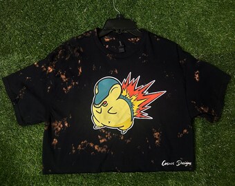 May include: Black cropped t-shirt with a bleach-splattered design. The shirt features a cartoon image of a yellow and blue Pokemon character with a red and orange flame tail. The text "Cosmos Designs" is printed on the bottom right corner of the shirt.