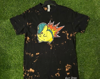 May include: Black t-shirt with a bleach-dye effect featuring a cartoon image of a yellow and orange Pokemon character with a green and blue hat. The text "Cosmos Designs" is printed on the bottom of the shirt.