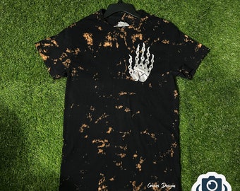 May include: Black t-shirt with a bleach-dye effect and a silver chain-link hand graphic on the chest. The shirt has short sleeves and the words "Cosmos Designs" printed at the bottom.