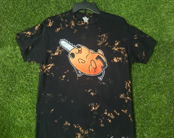May include: Black t-shirt with a bleach-dye effect featuring a cartoon orange character holding a chainsaw. The text "Cosmos Designs" is printed on the bottom right corner of the shirt.