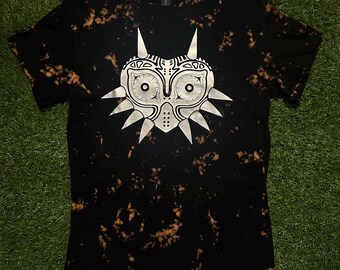 May include: A black t-shirt with a bleach-splattered design featuring a white silhouette of the Majora's Mask from the Legend of Zelda video game series. The text "Cosmos Designs" is printed in white at the bottom of the shirt.