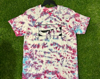 May include: A white tie-dye t-shirt with a black and white illustration of a character with a bandana and an eye patch. The character's name is written in Japanese below the illustration. The text "Cosmos Designs" is printed on the bottom right corner of the shirt.