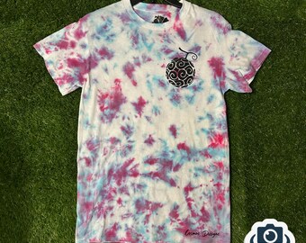 May include: A white tie-dye t-shirt with a black and white graphic of a fruit with a swirl design. The shirt has a blue and pink tie-dye pattern. The text "Cosmos Designs" is visible at the bottom of the shirt.