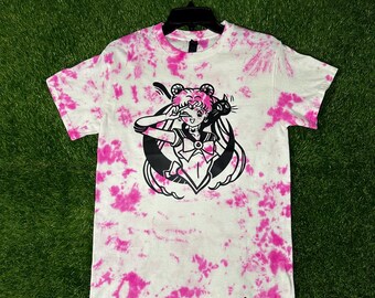 May include: A white t-shirt with a pink tie-dye pattern. The shirt features a black and white illustration of Sailor Moon with Luna, her black cat companion. The text "Cosmos Designs" is printed in black at the bottom of the shirt.