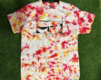 May include: A white tie-dye t-shirt with a black and white illustration of an anime character's eye with the Japanese word "ルフィ" (Luffy) below it. The shirt has a red, yellow, and white tie-dye pattern.