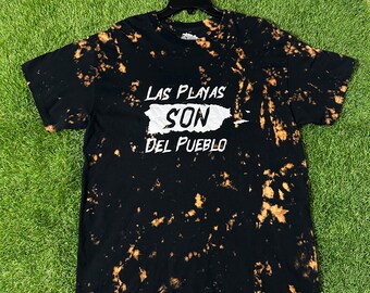 May include: Black t-shirt with a bleach tie-dye pattern. The shirt features a white graphic of the island of Puerto Rico with the text "Las Playas Son Del Pueblo" in white.