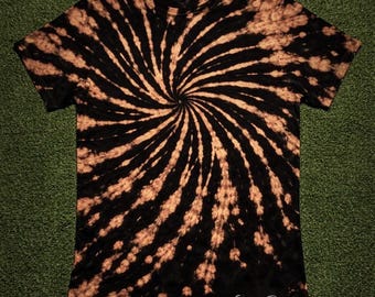 May include: Black t-shirt with a spiral tie-dye pattern in shades of brown and black. The design radiates from the center, creating a swirling effect. The text "Cosmos Designs" is visible at the bottom right.
