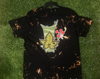 May include: Black t-shirt with a bleach-dye effect featuring a green cartoon creature with a red flower. The text "Cosmos Designs" is printed on the bottom right corner of the shirt.