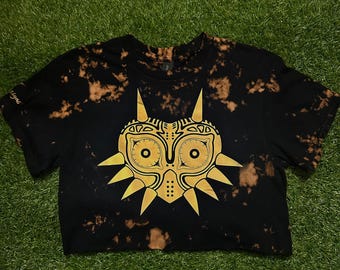 May include: Black crop top with a gold graphic of a mask. The shirt has a bleach-dye effect with brown spots. The mask design features eye-like circles and pointed edges. The shirt is laying on a green surface.