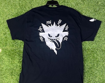 May include: Black t-shirt featuring a white graphic of a cartoon ghost with a long tongue and a menacing grin. The design is surrounded by stylized symbols. The shirt is hanging on a black hanger against a green background.