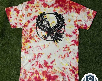 May include: White t-shirt with a tie-dye pattern in red and yellow hues. The back of the shirt features a black graphic of an eagle within a circular design. The shirt is short-sleeved and made of a soft material.