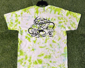 May include: White t-shirt with a tie-dye pattern in shades of green and blue. The shirt features a black graphic of a snarling creature with a snake wrapped around it. The shirt is hanging on a black hanger.