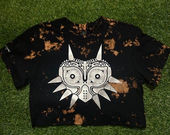 May include: Black crop top with a bleach-dye effect and a silver graphic of a mask. The mask design features sharp points and circular eye details. The shirt is displayed on a green surface.