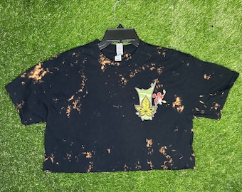 May include: Black cropped t-shirt with a bleach tie-dye pattern. The shirt features a cartoon character with green leaves and a red flower.