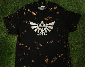 May include: Black t-shirt with a white bleach tie-dye pattern and a silver Triforce symbol from the Legend of Zelda video game series. The shirt has the text "Cosmos Designs" on the tag.