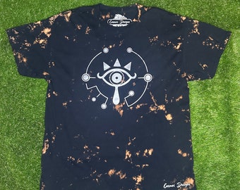 May include: Black t-shirt with a bleach-dye effect and a silver graphic of the Sheikah Eye from The Legend of Zelda: Breath of the Wild. The shirt has the text "Cosmos Designs" on the tag.