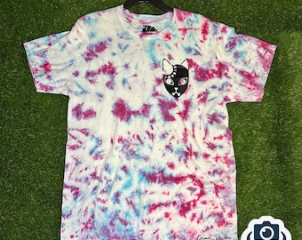 May include: A white tie-dye t-shirt with a black cat face design on the front. The shirt has a blue and pink tie-dye pattern. The text "Cosmos Designs" is printed on the bottom of the shirt.