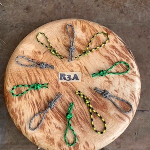 Könnte beinhalten: Acht verschiedene farbige Paracord-Schlaufen mit Knoten auf einer Holzoberfläche. Die Schlaufen sind in verschiedenen Farben, darunter Grün, Gelb, Schwarz und Grau. Der Text "R3A" ist auf einem weißen Etikett in der Mitte der Holzoberfläche geschrieben.