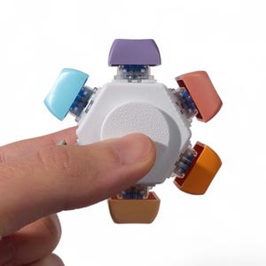 May include: A multi-colored fidget toy with a white hexagonal body and six colored buttons. The buttons are light blue, orange, peach, purple, and tan. The toy is held in a person's hand against a white background.