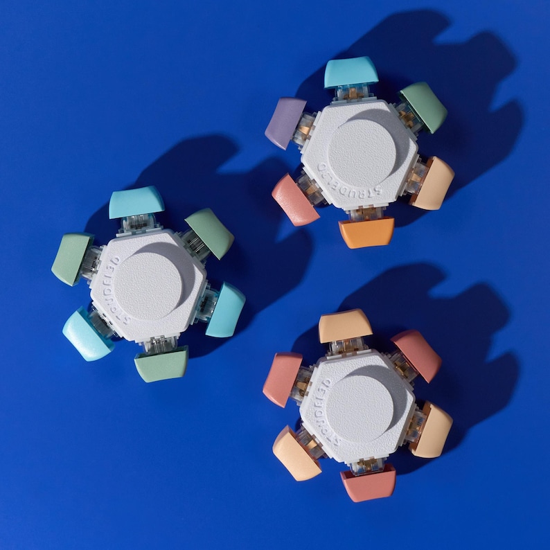Puede incluir: Tres juguetes fidget blancos con botones de colores pastel, cada uno con la palabra "STRUDEL" impresa. Cada juguete tiene un centro hexagonal con seis extensiones coloridas en tonos de azul, verde, naranja, rosa y morado. El fondo es azul liso.