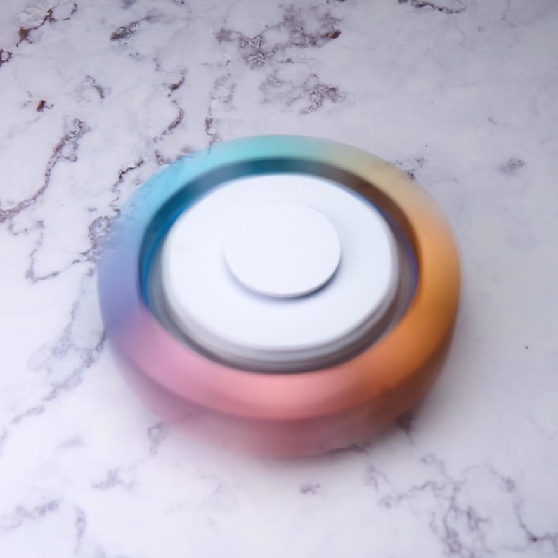 May include: A white and rainbow-colored fidget spinner. The spinner has a white center and a colorful outer ring that blends from blue to pink to orange. The background is a marble-like surface.