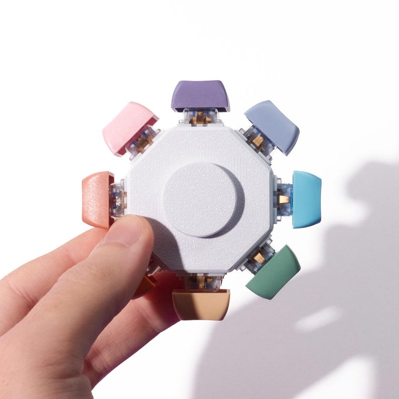 May include: A multi-colored fidget toy with a white central hub and eight pastel-colored buttons. The buttons are pink, peach, purple, blue, green, and tan. The toy is held in a hand against a white background.