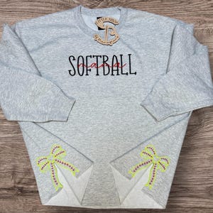 May include: A light gray sweatshirt with the word "SOFTBALL" embroidered in black and red. The sleeves feature softball-themed bow designs in yellow, pink, and green. A wooden tag with text is attached to the neckline.