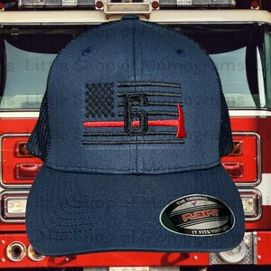 May include: A navy blue baseball cap with a black and red American flag design. The flag has a red line across it and the number "6" embroidered on it. The cap has a black mesh back and a Flexfit label.