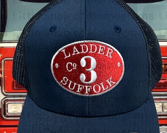 May include: Navy blue Flexfit hat with a mesh back. The front features a red oval patch with white text that reads "LADDER Co 3 SUFFOLK". The hat has a curved brim and the Flexfit logo.