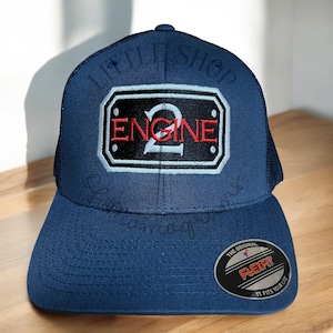May include: A navy blue trucker hat with a black and white patch that says "Engine 2" in red lettering. The hat has a Flexfit label on the side.