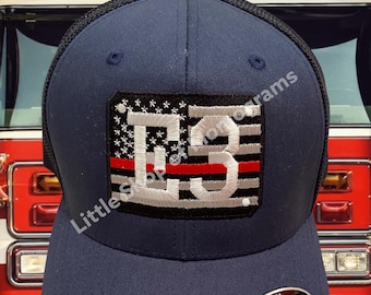 Può includere: Un cappello da baseball blu navy con retro in rete nera. Il cappello presenta una bandiera americana ricamata in bianco con una striscia rossa al centro e il numero "E3" in bianco.