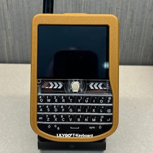 May include: A gold-colored handheld device with a black screen and a black keyboard. The keyboard has the letters QWERTYUIOP and numbers 123. The device has the text "LILYGO® T-Keyboard" on the bottom.