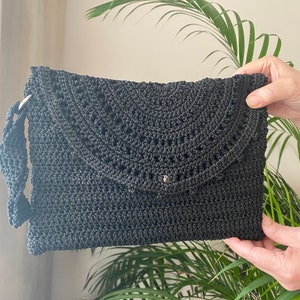 Puede incluir: Una cartera de mano negra de ganchillo con un diseño decorativo de calado. La bolsa tiene una sola correa.