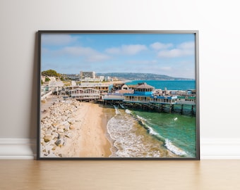 May include: An aerial view of a pier with several restaurants and shops on it. The pier is located on a sandy beach with blue water and white waves. There are buildings in the background.