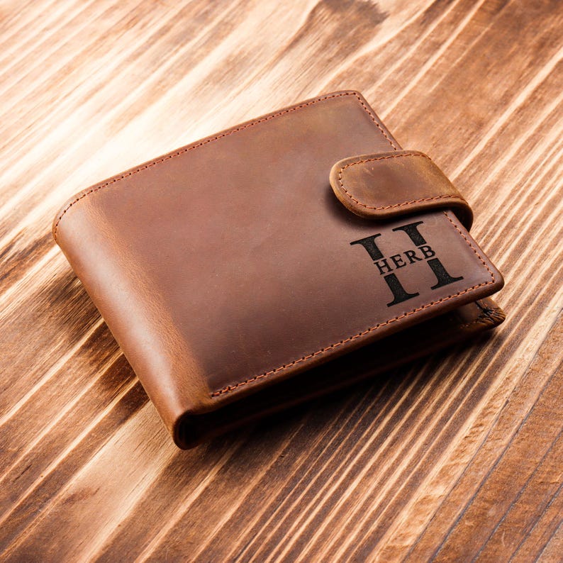 Puede incluir: Una cartera de cuero marr&oacute;n con cierre de solapa y un monograma personalizado que dice "II HERB II".