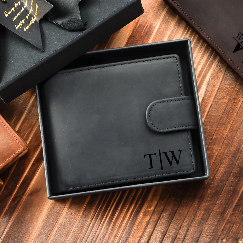 Puede incluir: Cartera de cuero negro con cierre de solapa y un monograma personalizado "T|W" en una caja negra.