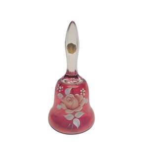 May include: A ruby red glass bell with a clear handle. The bell features a hand-painted design of a pink rose with white and green accents. A gold sticker is on the handle. Decorative item.