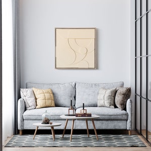 May include: A light gray sofa with three pillows, two coffee tables, and a beige abstract art print on the wall. The sofa is in a living room with a window and a glass door.