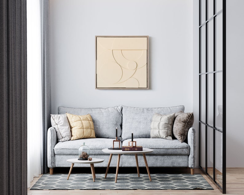 May include: A light gray sofa with three pillows, two coffee tables, and a beige abstract art print on the wall. The sofa is in a living room with a window and a glass door.