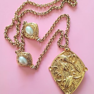 Op de afbeelding: Goudkleurige ketting en oorbellen set. De ketting heeft een grote, sierlijke hanger met een bloemenontwerp en een ketting. De oorbellen hebben een parelachtige steen. De sieraden staan tegen een roze achtergrond.