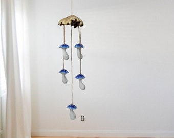 May include: A wooden mushroom wind chime with five blue and white ceramic mushrooms hanging from it. The wind chime is hanging from the ceiling in a room with white walls and wooden floors.
