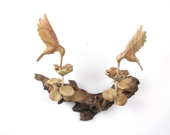 May include: Two wooden hummingbirds carved in detail, perched on a driftwood branch with carved flowers. The birds are in flight, with their beaks extended as if feeding from the flowers.