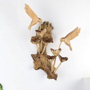May include: Wooden wall art featuring two carved hummingbirds with outstretched wings, feeding from mushroom-shaped flowers. The sculpture is mounted on a natural wood base, showcasing the grain and texture of the wood. The piece is a light brown color.