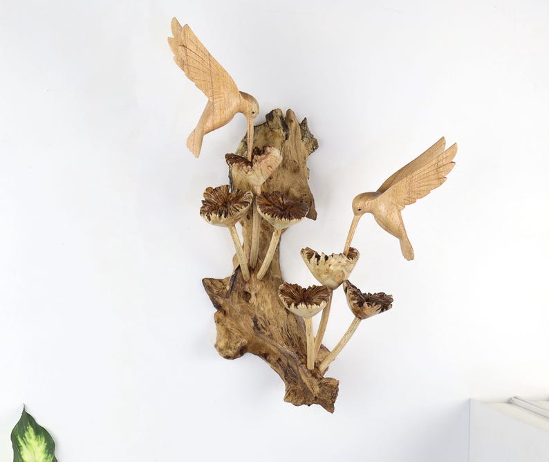 May include: Wooden wall art featuring two carved hummingbirds with outstretched wings, feeding from mushroom-shaped flowers. The sculpture is mounted on a natural wood base, showcasing the grain and texture of the wood. The piece is a light brown color.