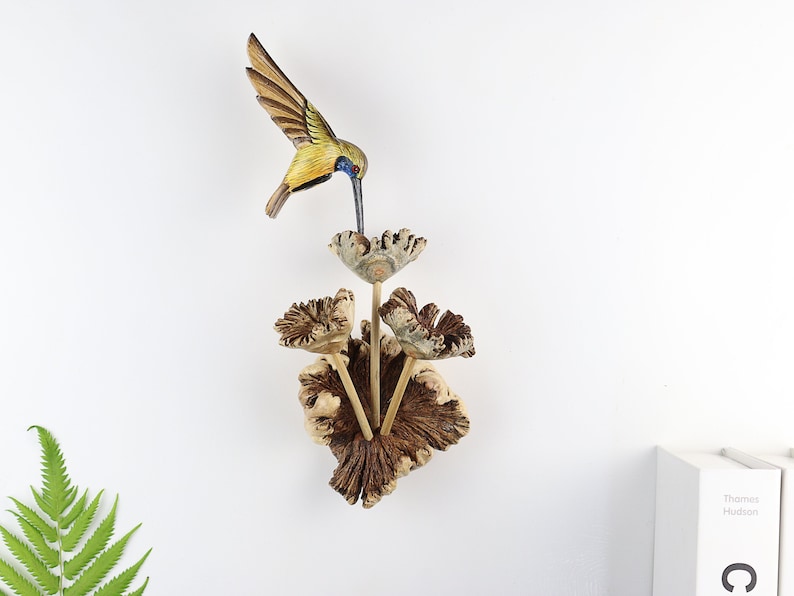 K&ouml;nnte beinhalten: Ein handgeschnitzter Holz-Kolibri-Futterautomat mit einem detaillierten Kolibri im Flug, der sich von einem bl&uuml;tenf&ouml;rmigen Reservoir ern&auml;hrt. Der Futterautomat ist aus Naturholz gefertigt und hat gelbe, gr&uuml;ne und blaue Akzente.