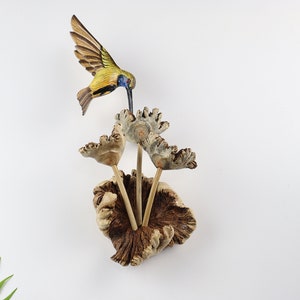 K&ouml;nnte beinhalten: Eine Holzskulptur eines Kolibris mit ausgebreiteten Fl&uuml;geln, der &uuml;ber einer Gruppe stilisierter Holzblumen schwebt. Der Kolibri hat gelbe, blaue und braune Farben. Die Blumen sind hellbraun und die Basis ist dunkler braun.