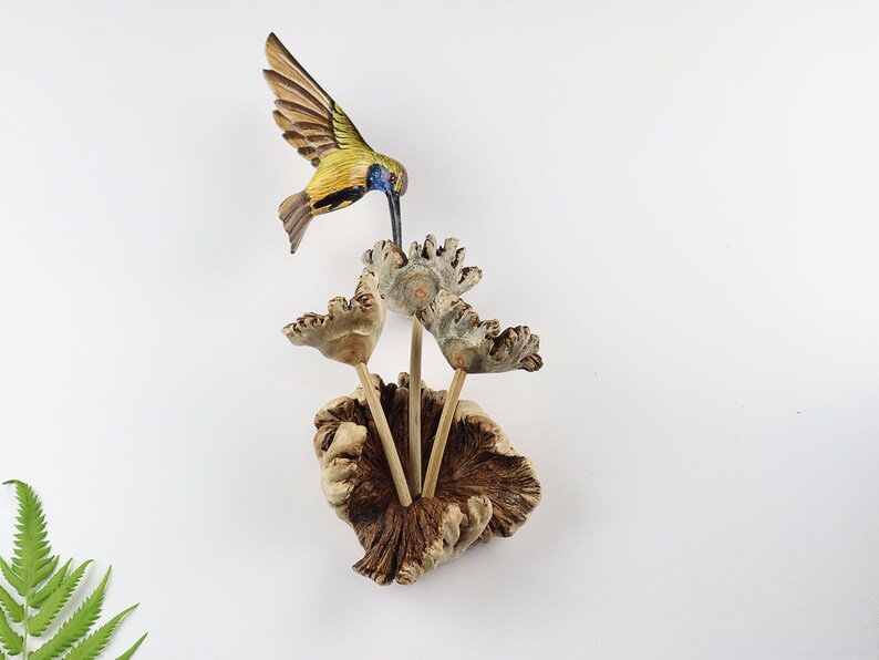 K&ouml;nnte beinhalten: Eine Holzskulptur eines Kolibris mit ausgebreiteten Fl&uuml;geln, der &uuml;ber einer Gruppe stilisierter Holzblumen schwebt. Der Kolibri hat gelbe, blaue und braune Farben. Die Blumen sind hellbraun und die Basis ist dunkler braun.