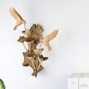 May include: A wooden wall sculpture featuring two hummingbirds and stylized flowers. The birds are carved in detail, with outstretched wings and long beaks. The sculpture is mounted on a piece of textured wood, creating a natural and artistic display.