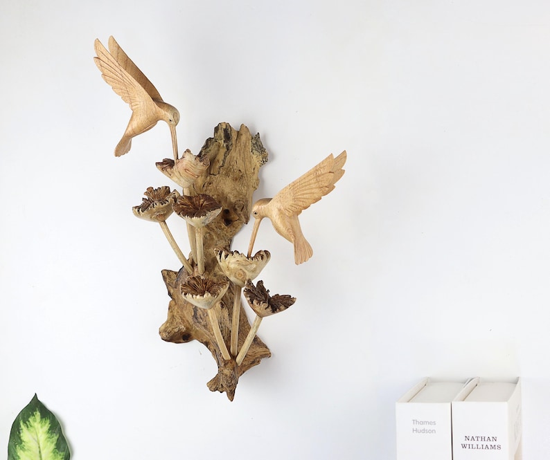 May include: A wooden wall sculpture featuring two hummingbirds and stylized flowers. The birds are carved in detail, with outstretched wings and long beaks. The sculpture is mounted on a piece of textured wood, creating a natural and artistic display.